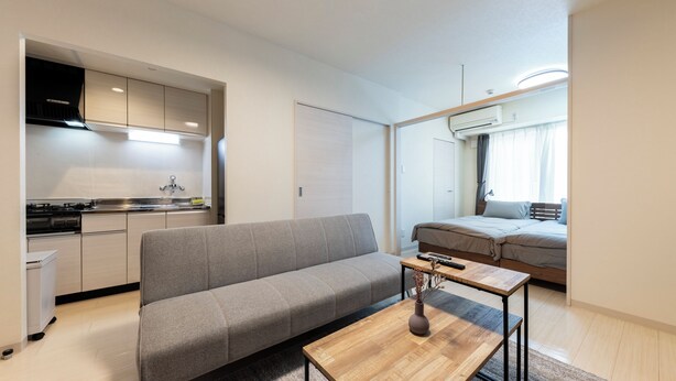Un appartement moderne avec un canapé gris, une table basse en bois, une cuisine équipée d’une cuisinière et un lit pr ès d’une fenêtre.