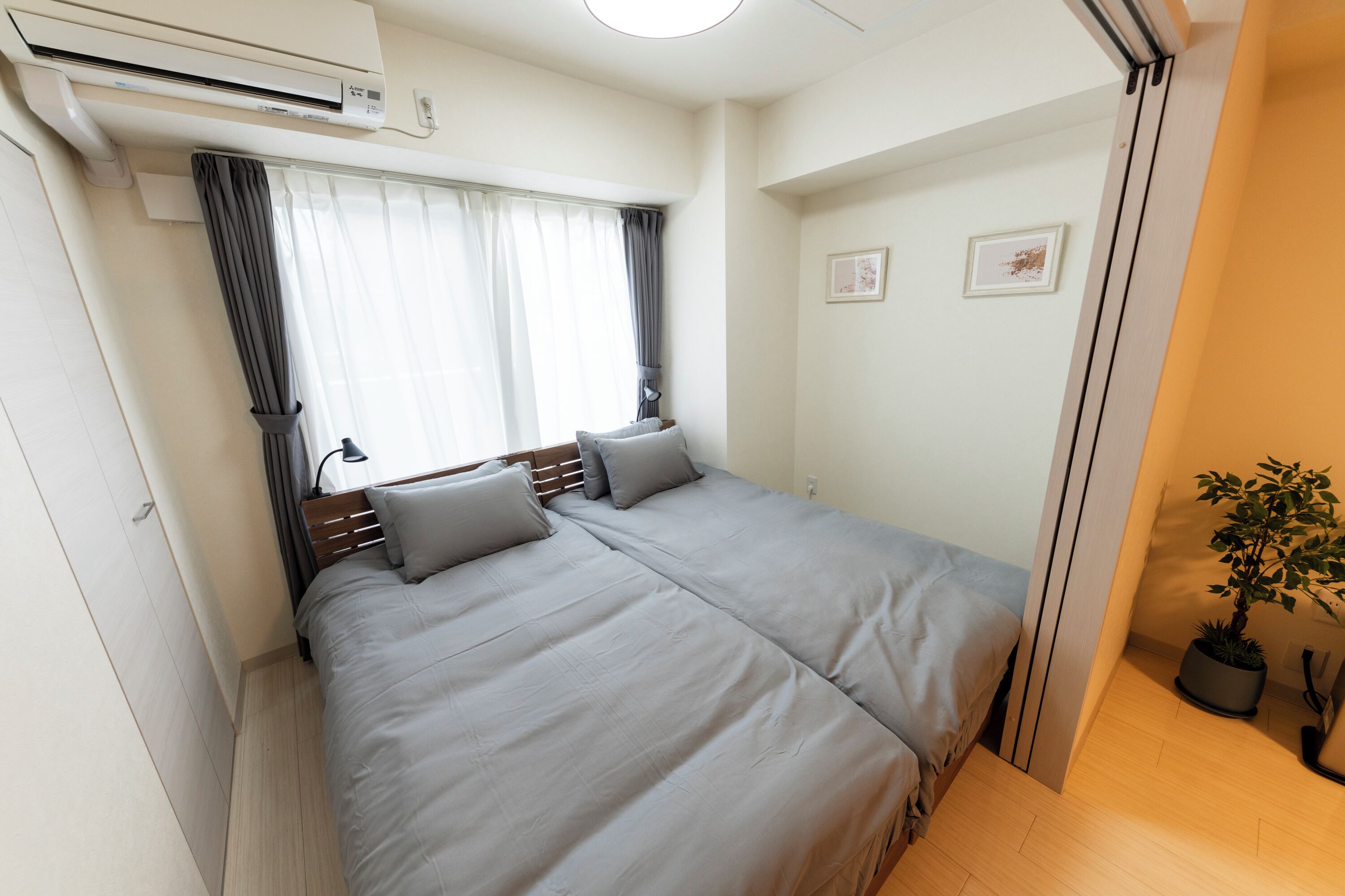 A bedroom with a bed, air conditioning unit, curtains, a potted plant, and a small framed picture on the wall.