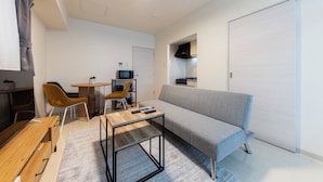 Un salon moderne avec un canapé gris, une table basse en bois et deux fauteuils. On aperçoit à l’arrière-plan un coin cuisine avec une cuisinière et des placards.