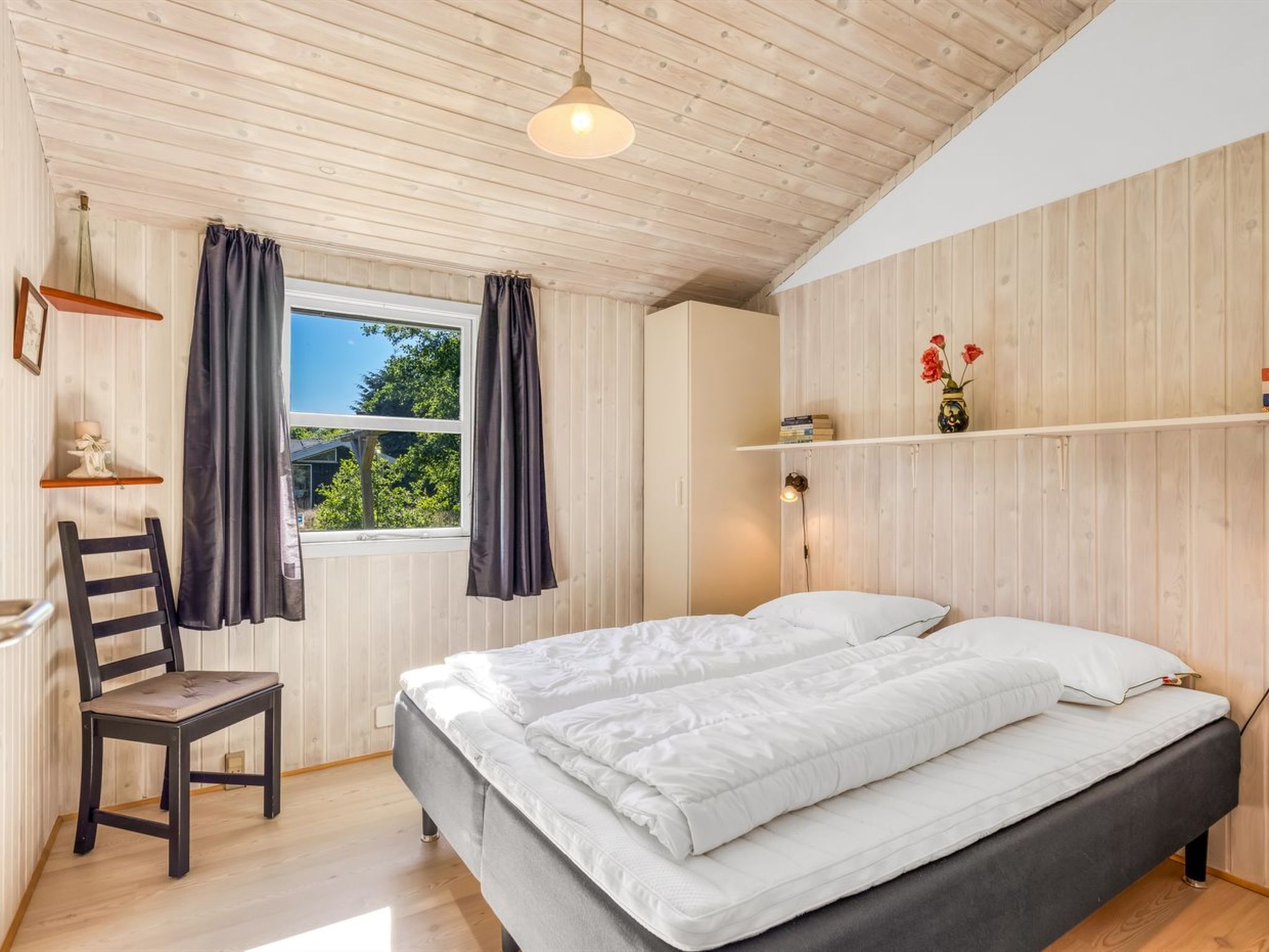 Ein Schlafzimmer mit einem Bett, einem Stuhl, einem Fenster mit Vorhängen, einem Regal mit Büchern und Blumen sowie einer Holzbalkendecke.