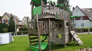 Eine Holzspielanlage mit Rutsche und Kletterelementen, eingebettet in eine Rasenfläche, im Hintergrund sind Häuser zu sehen.