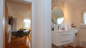 Ein modernes Hotelzimmer mit Holzboden, einer Essecke mit Tisch und Stühlen, einem weißen Schminktisch mit Spiegel und einem Fenster mit Vorhängen.