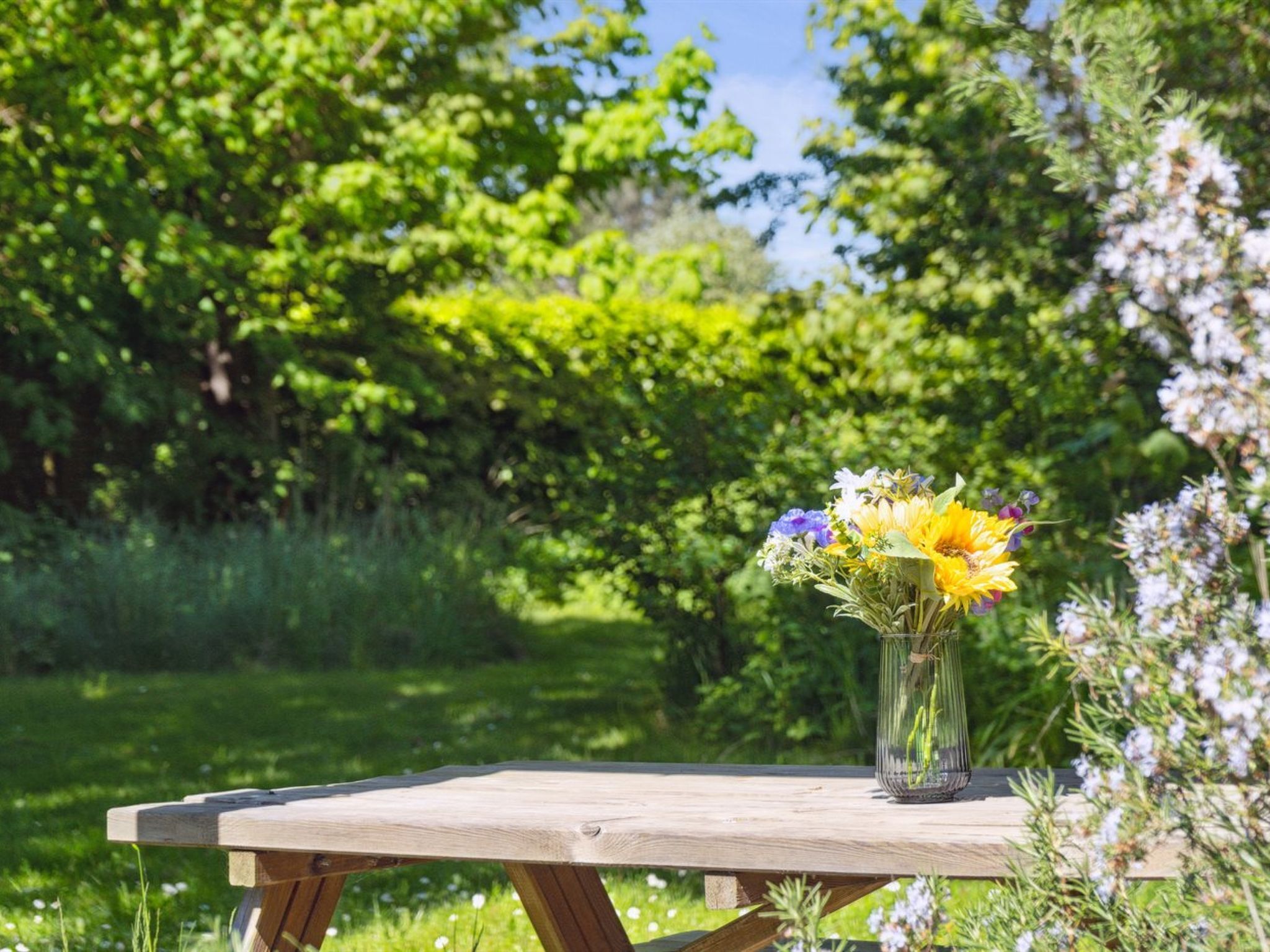 Ein Holztisch mit einer klaren Glasvase mit Blumen, draußen aufgestellt, mit üppigem Grün im Hintergrund.