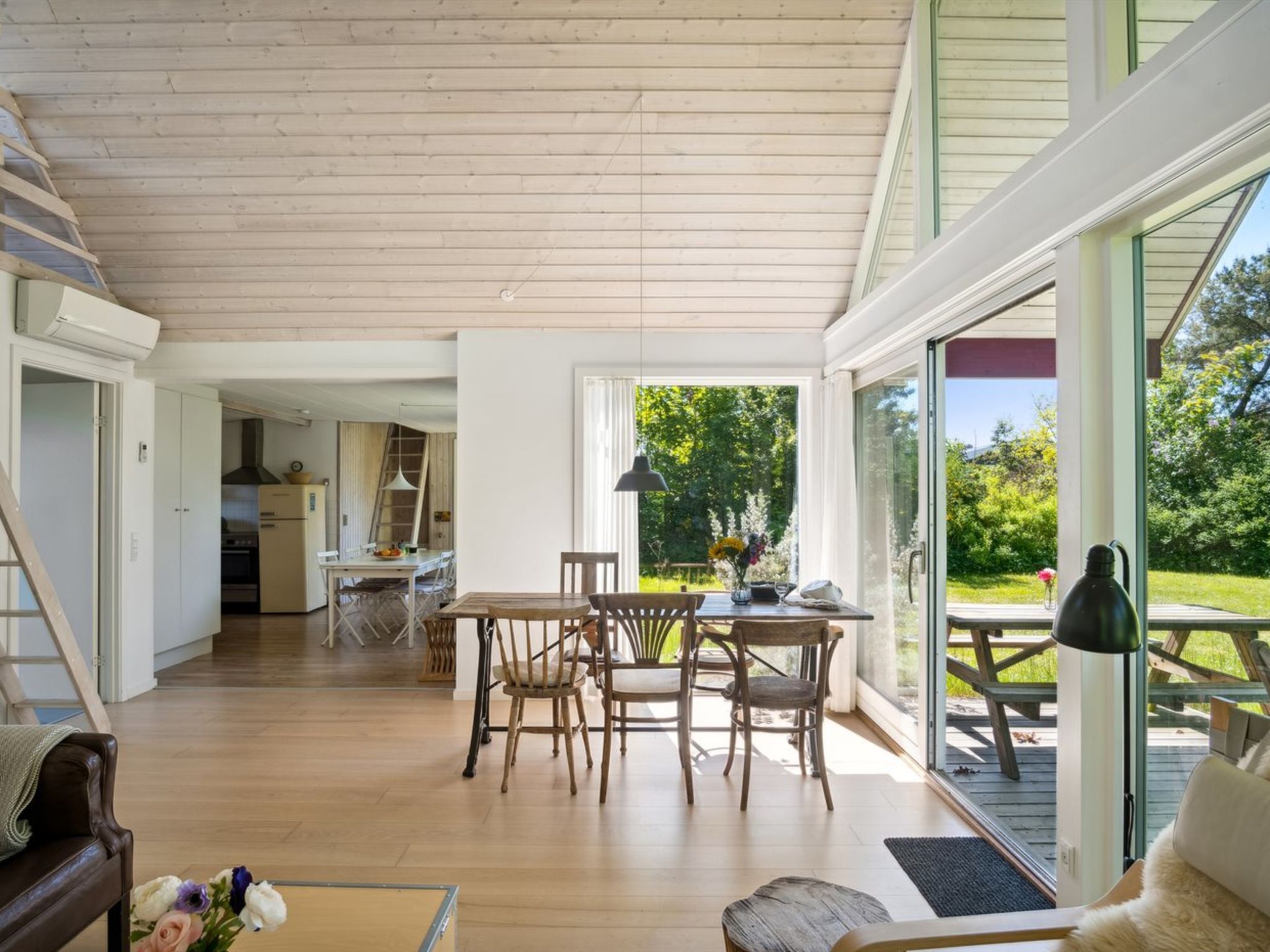 A spacious living area with a wooden floor, a dining table with chairs, a sofa, and a view of the outdoors through large windows.