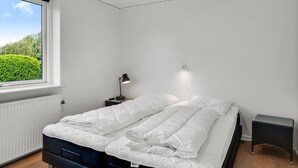 Un dormitorio con un ventanal grande, una cama con ropa de cama blanca, una mesita de noche con una lámpara y una pequeña mesita auxiliar.