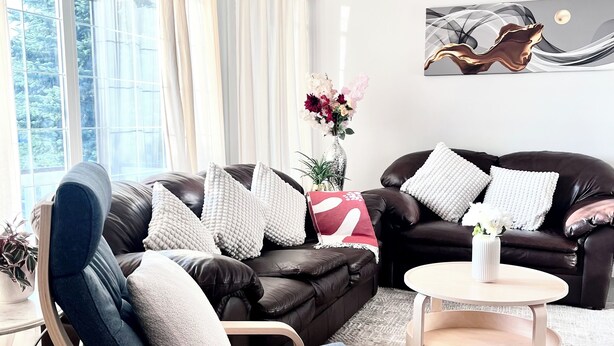 A modern living room with a leather sofa, a wooden chair, a round coffee table, and a large window with curtains. - Bungalow | Upper Level | CT Train | Spruce Meadow (Calgary)