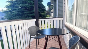A balcony with a round table and two chairs, a white picket fence, and a view of a house and trees. - Bungalow | Upper Level | CT Train | Spruce Meadow (Calgary)