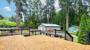 A wooden deck with a black railing, surrounded by a forested area with tall trees and a small shed. - A Cozy HomeAway Home @Bridle Trail, Bellevue, WA (Bellevue)