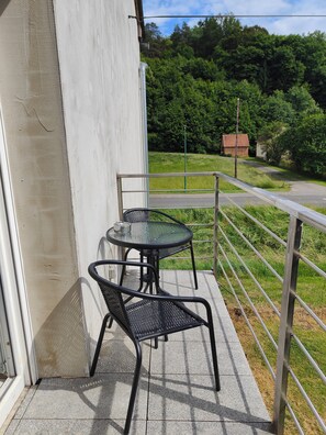 A balcony with a metal table and two chairs, overlooking a road and greenery.