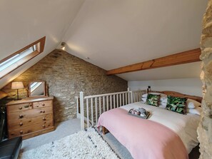 A bedroom with a stone wall, wooden beams, a dresser, a bed with a pink blanket, and a small table with cups.