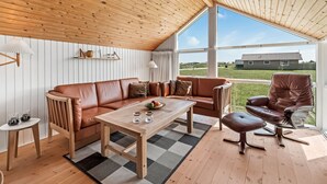 Een gezellige woonkamer met een houten plafond, een bruine leren bank, een houten salontafel en uitzicht op een huis door grote ramen.