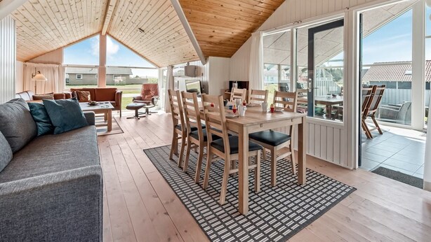 Een ruime kamer met een hoge plafond, houten balken, een eethoek met stoelen en een tafel, een bank en grote ramen met uitzicht op het omliggende landschap.