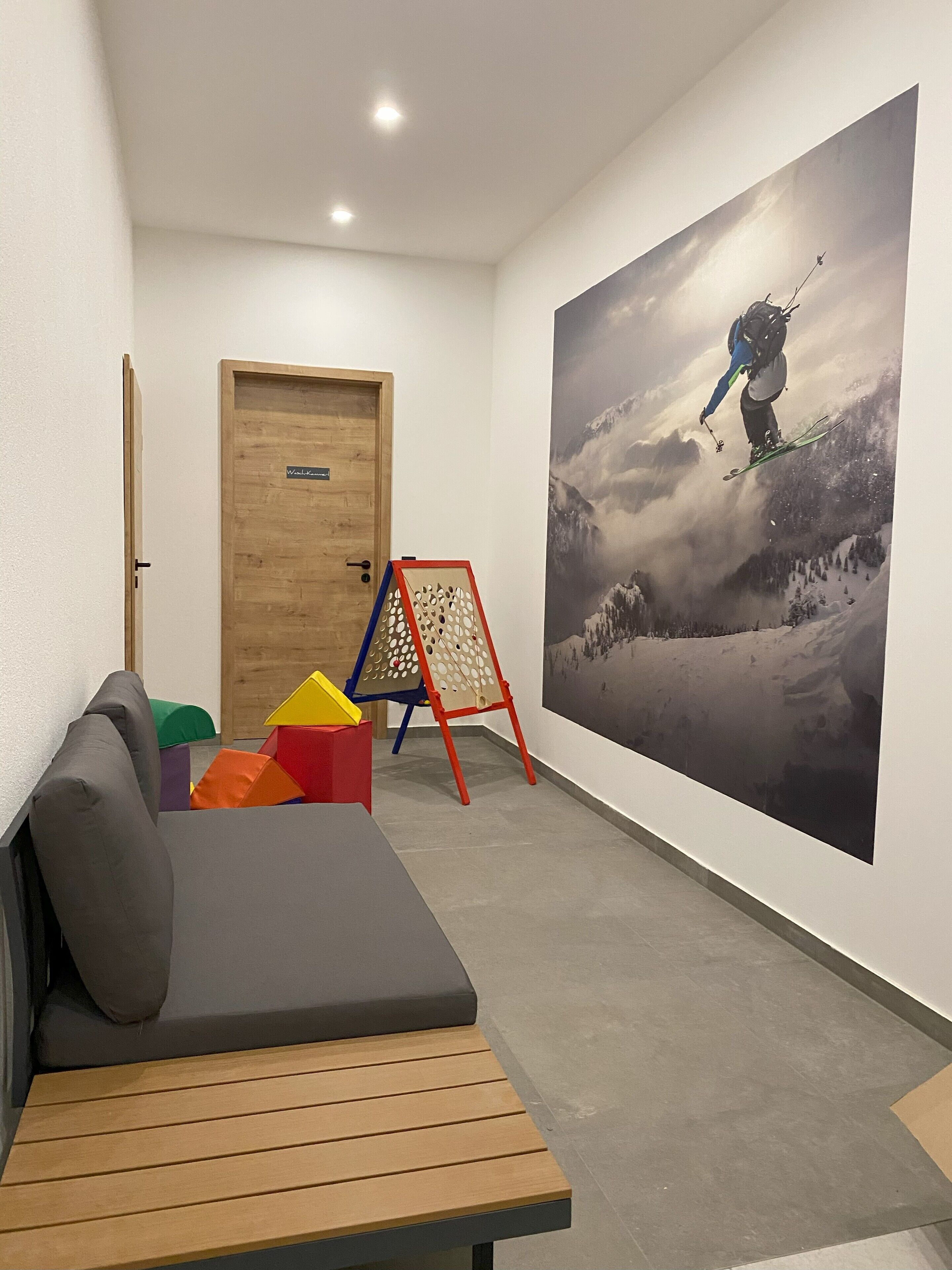 Un couloir avec une porte en bois, une structure de jeu colorée, une grande fresque murale représentant un skieur et un banc.