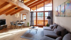 Un salon spacieux avec un plafond en bois, une grande fenêtre, un canapé gris, un coin repas avec des chaises et un téléviseur à écran plat.
