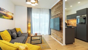 Un salon moderne avec un canapé gris, des coussins jaunes, une table basse et un téléviseur. La pièce est dotée d’un plancher en bois et d’une grande fenêtre avec des rideaux.