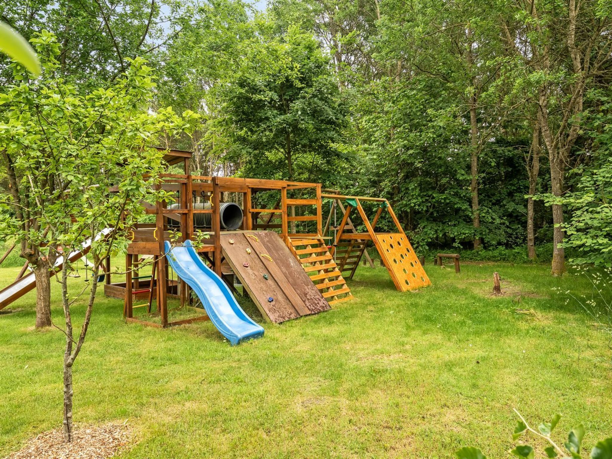 Ein hölzerner Spielplatz mit Rutsche, Kletternetz und einem kleinen Baum.
