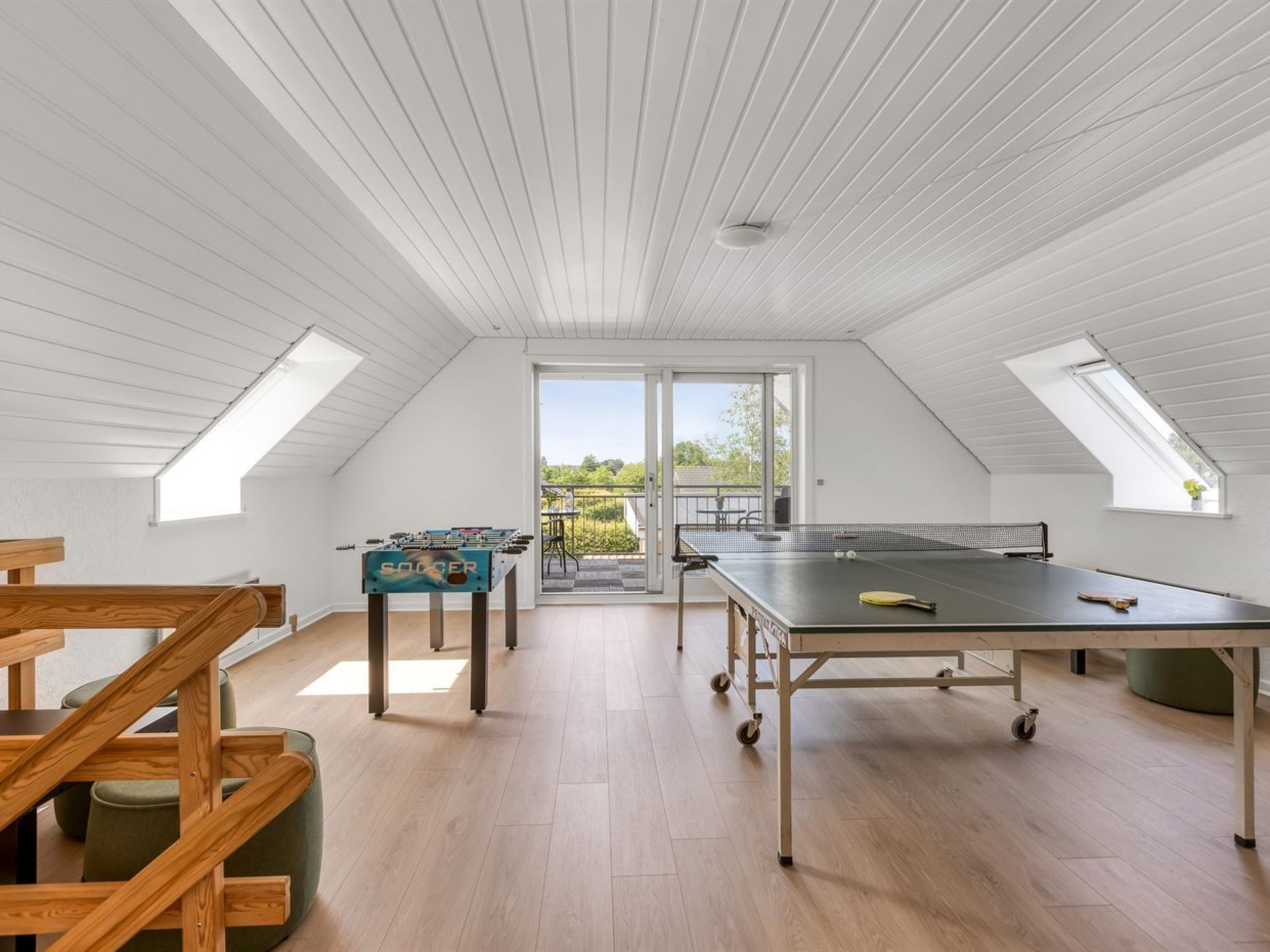 Una habitación amplia con mesa de ping pong, mesa de futbolín y una escalera con barandillas de madera.