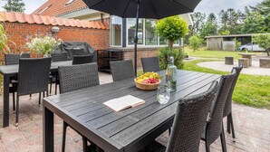 En udendørs terrasse med et træbord, fletstole, en grill og en stor parasol.