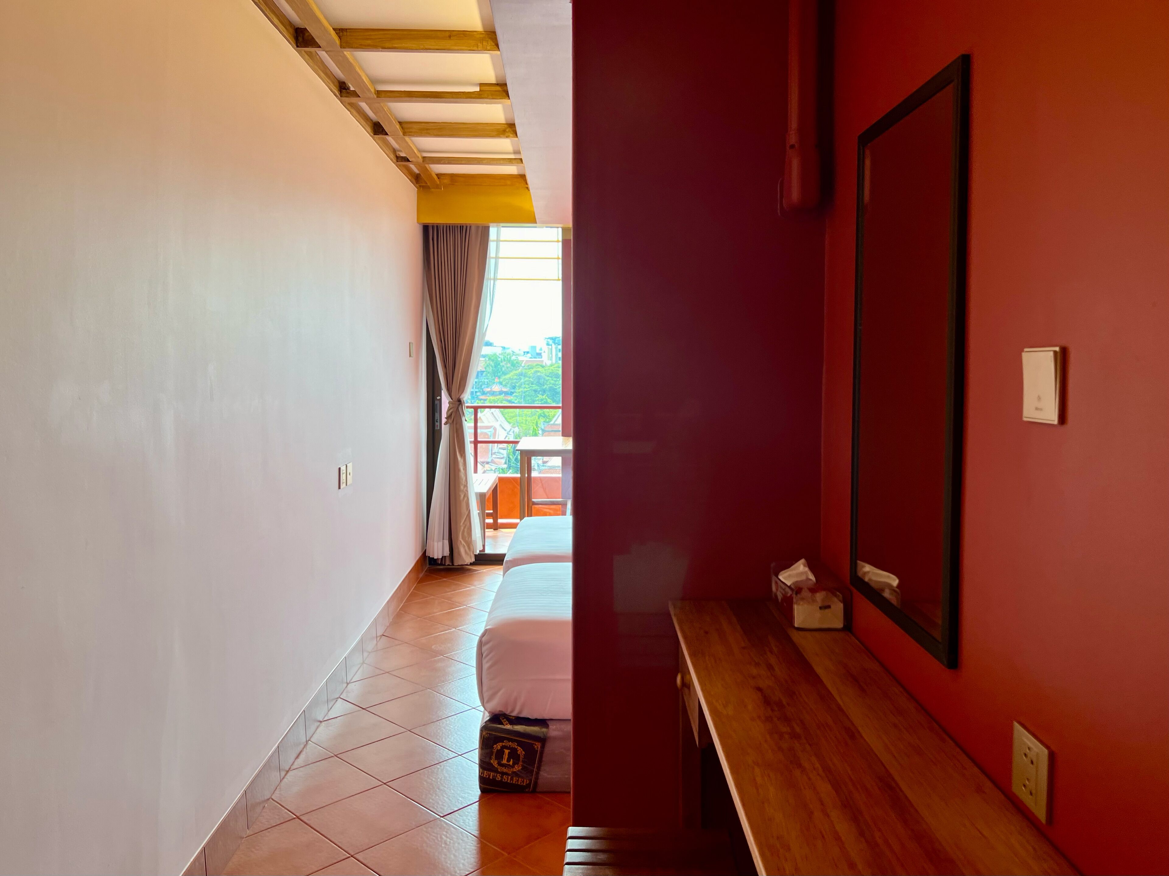 Habitación de hotel con cama, banco de madera, pared roja, ventana con cortinas y vista a la vegetación.
