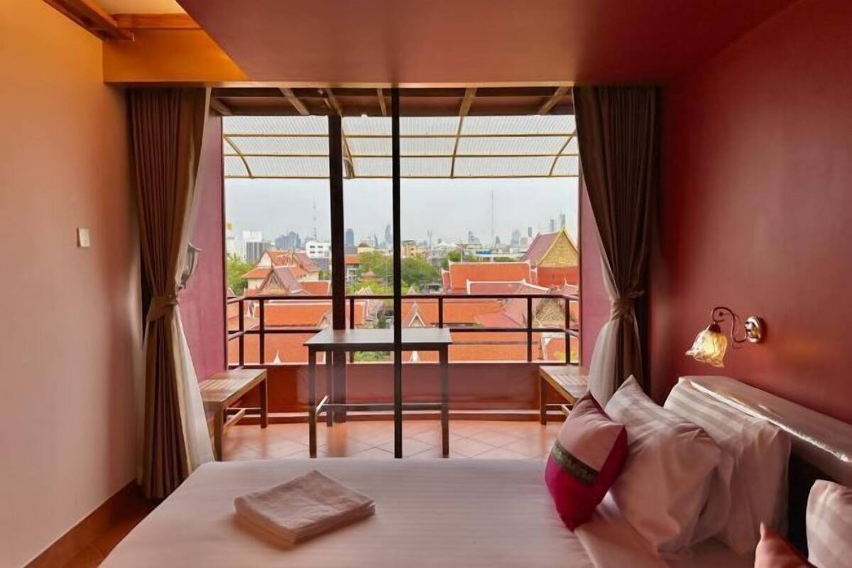 A hotel room with a bed, a small table, a bench, and a view of the cityscape through large windows.