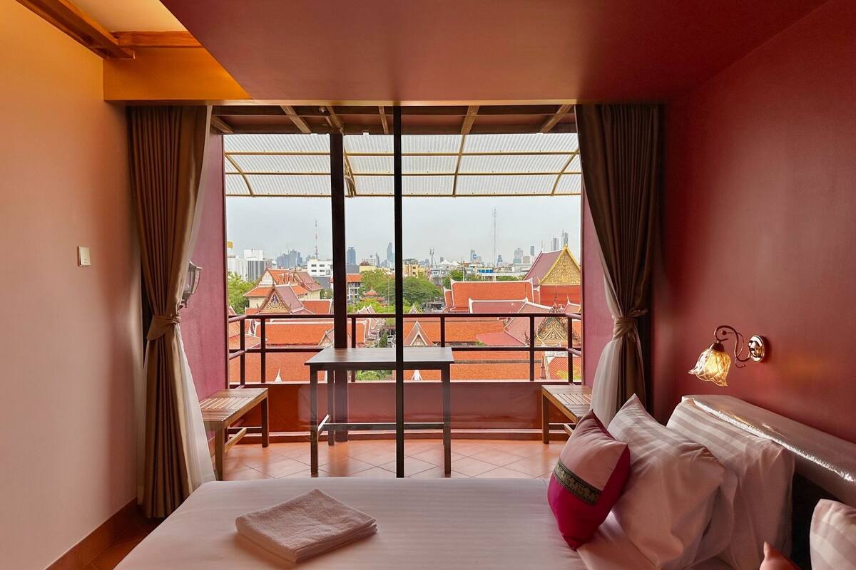 A hotel room with a bed, a small table, and a view of buildings through large windows.