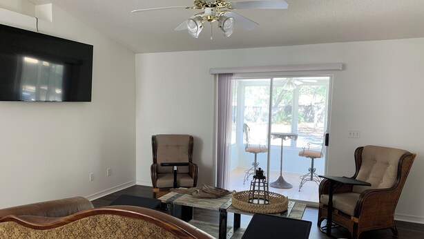 A living room with a sofa, two armchairs, a coffee table, and a television mounted on the wall. There is a ceiling fan and a view of the outdoors through a sliding glass door. - Newly Renovated, 1/2 acre home 3 miles from beach (Navarre)