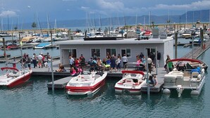 Un puerto deportivo con varias embarcaciones amarradas, una estructura cubierta con gente en la cubierta, y una vista despejada del agua y montañas distantes.