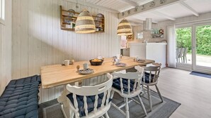 Een eetgedeelte met een houten tafel, witte stoelen en een bank. Boven de tafel hangen twee lampen.