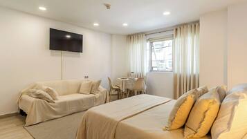 A hotel room with a bed, a sofa, a TV mounted on the wall, a dining table with chairs, and a window with curtains.