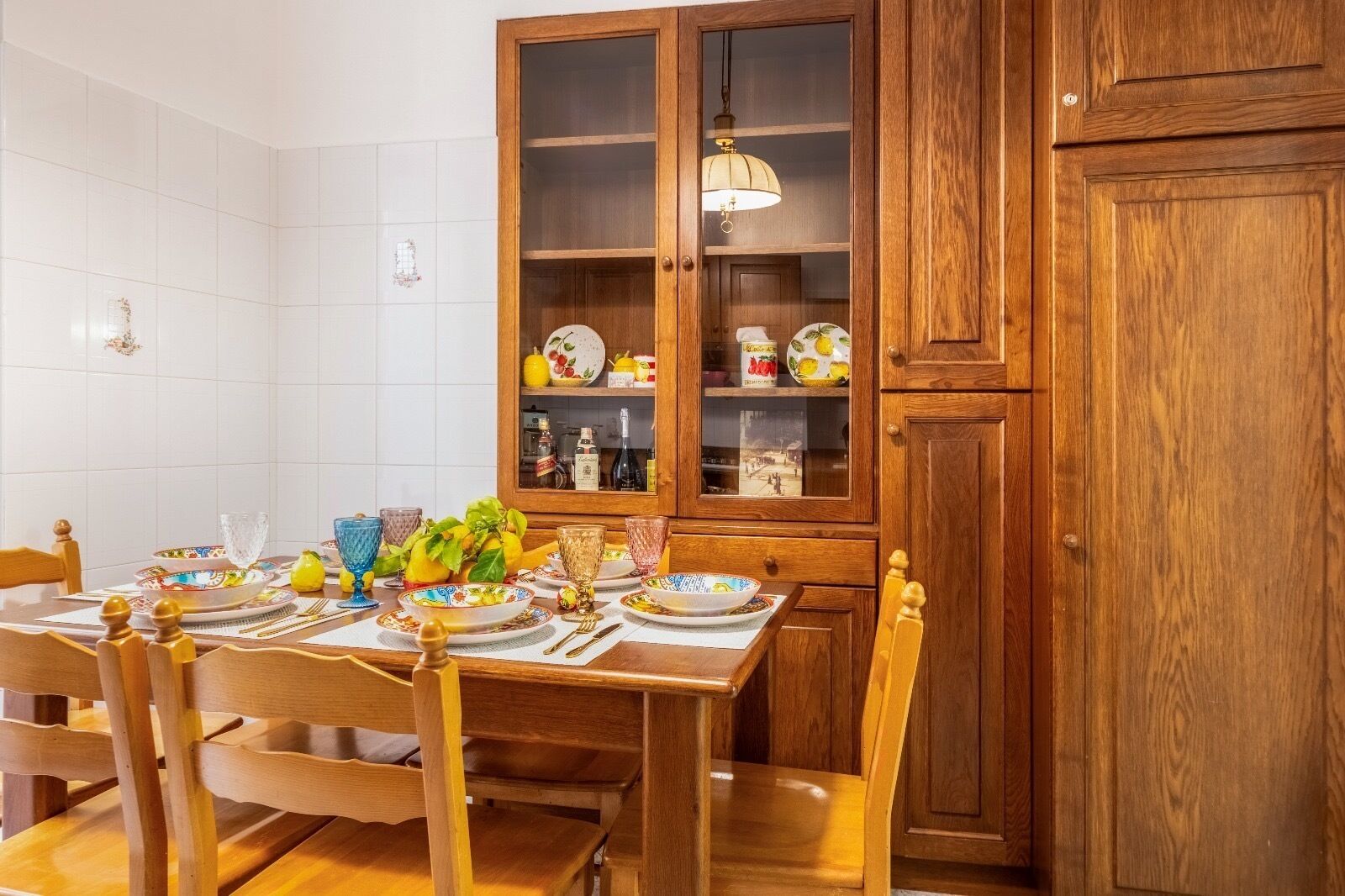 A dining area with a wooden table set for four, surrounded by wooden chairs. A wooden cabinet with glass doors is visible in the background.