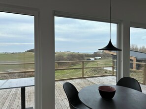 A room with a large window overlooking a rural landscape, a round table with chairs, and a hanging light fixture. - Cozy tiny house in nature - Holiday home D (Sejerø)