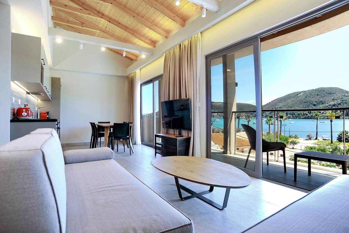 A modern living room with a sofa, a dining table, and a TV. There is a balcony with a view of the sea and a mountain.