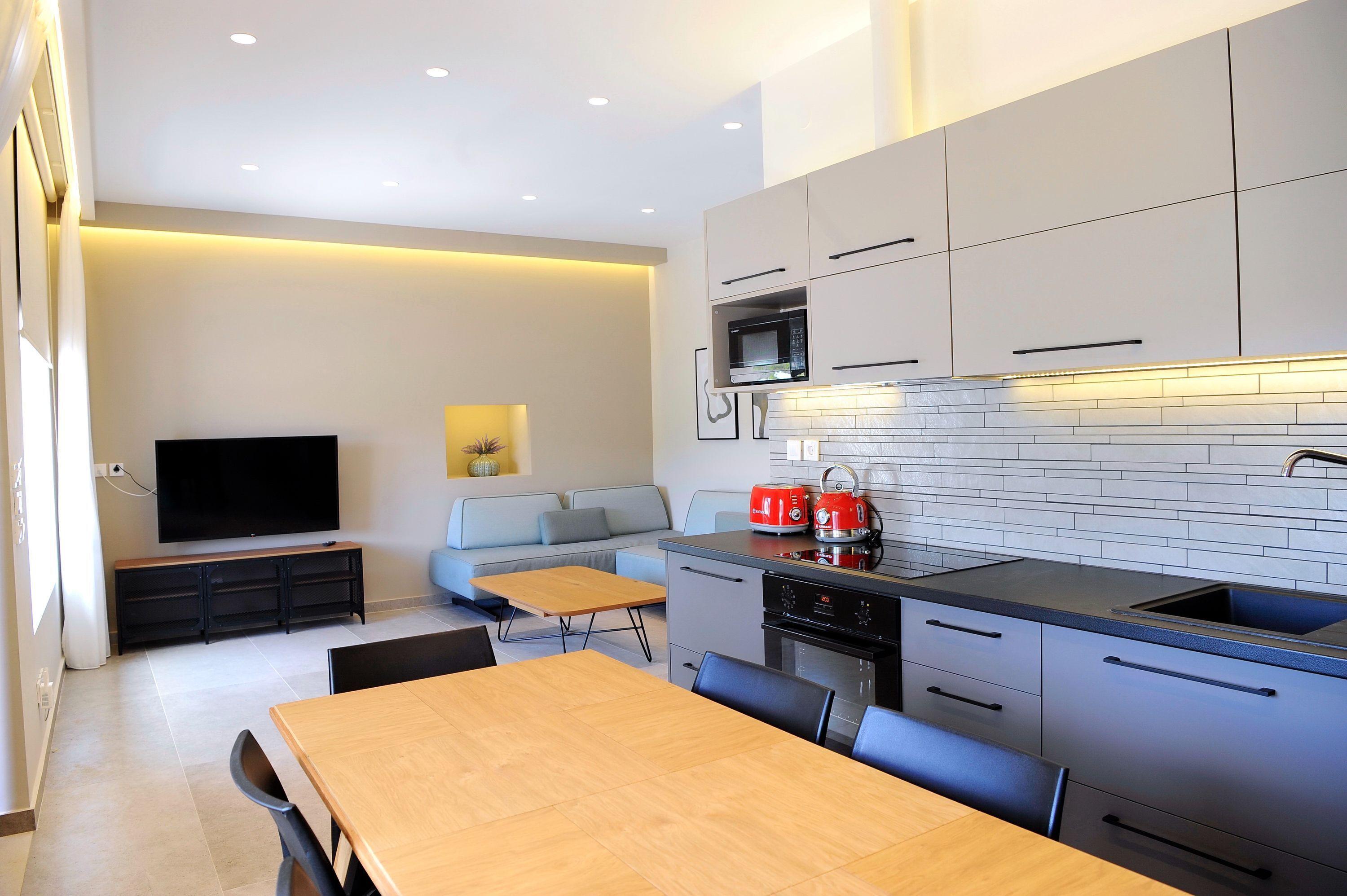 Une cuisine moderne avec des meubles gris, un four à micro-ondes et un évier. On y trouve une table à manger avec des chaises, et un canapé à l'arrière-plan.