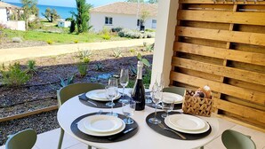 Un espace de restauration en plein air offrant une vue sur la mer, aménagé avec quatre chaises vertes et des tables rondes blanches, chacune étant dressée avec des assiettes, des couverts et des verres à vin.