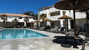 Un espace piscine agrémenté de chaises longues et de parasols en chaume, entouré d’un bâtiment doté de balcons, le tout sous un ciel d’un bleu limpide.