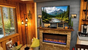 A cabin-style room with a stone fireplace, a television mounted above it, and a view of a mountain landscape. - Romantic Small Cabin in the Woods. Fireplace,Hot tub,A/C,Firepit,WiFi, Mt Rainer (Ashford)
