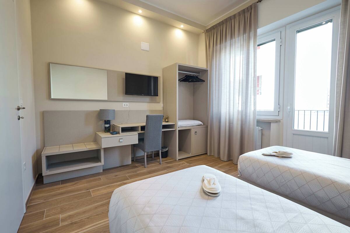 A modern hotel room with two beds, a desk, a chair, a TV, and a wardrobe.