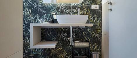 A bathroom with a modern design, featuring a large mirror, a white sink, and a decorative leaf-patterned wall.