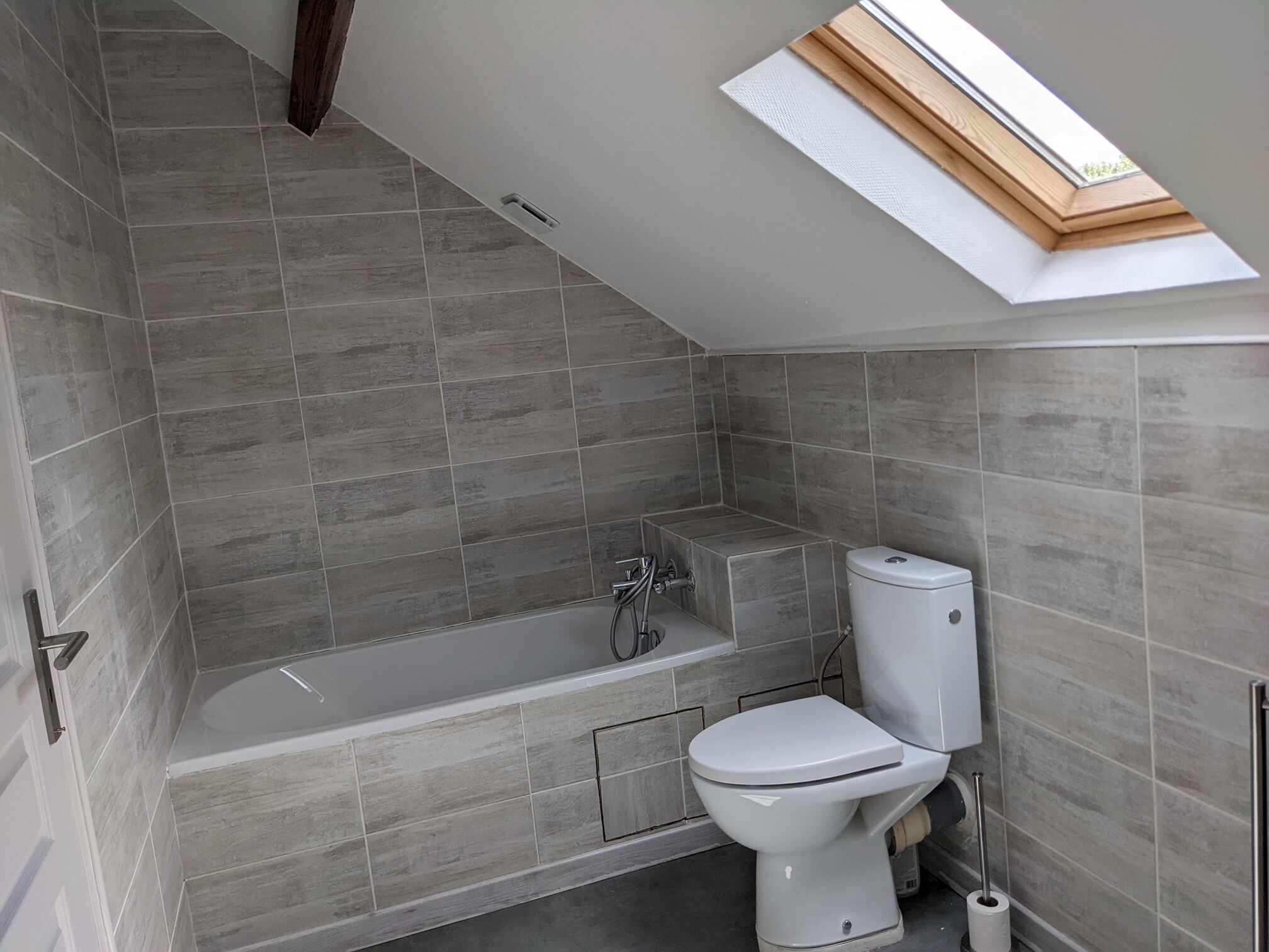 A modern bathroom with grey tiles, a bathtub, a toilet, and a skylight.