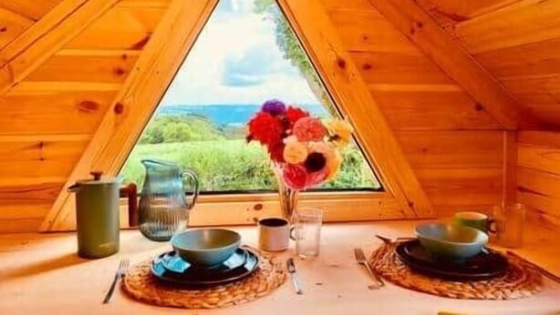 A wooden interior with a triangular window, a table set for two with plates, bowls, cups, and a pitcher. - Stairway to Devon - Tarka Cabin (Umberleigh)