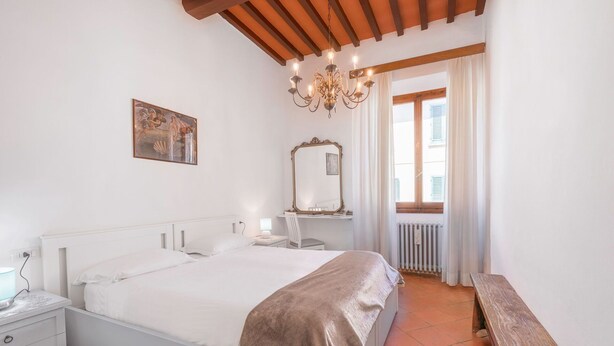 Ein Schlafzimmer mit einem großen Bett, einem Nachttisch, einem Sessel, einer Bank, einem Spiegel und einem Fenster mit Vorhängen.