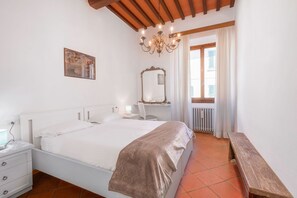 Une chambre à coucher avec un grand lit, une table de chevet, une chaise, un banc, un miroir et une fenêtre avec des rideaux.