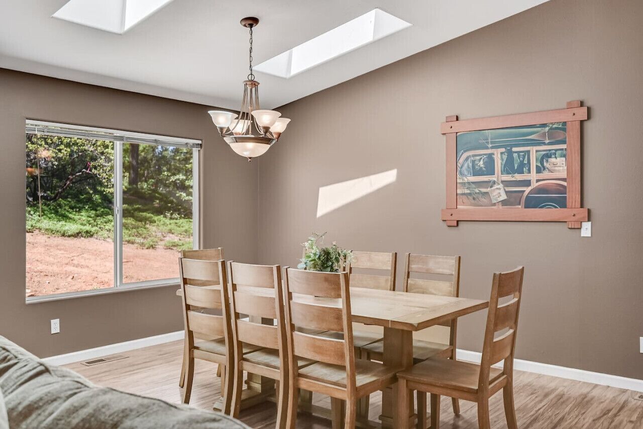 A dining area with a wooden table and chairs, a chandelier, a large window with a view of greenery, and a framed picture on the wall.