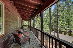 Une véranda en bois, dotée d’une balustrade, aménagée avec des fauteuils et une table en osier, et offrant une vue sur une zone boisée.
