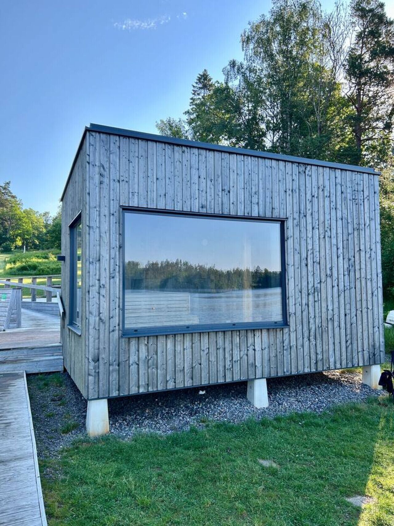 A modern wooden cabin with a large window, situated on a grassy area with a wooden deck.