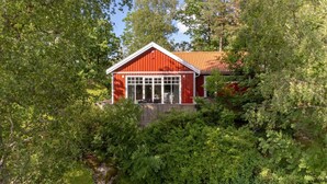 A red wooden building with a large window and a porch, surrounded by dense green foliage. - Lakeview spacious family home (Västra Götalands län)