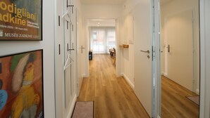 Un pasillo con piso de madera, paredes blancas y una puerta que da a una habitación. Hay un cartel en la pared y un felpudo en la entrada.