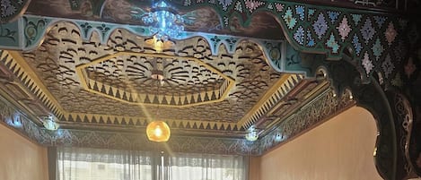 Una habitación con suelo de madera, dos sofás con cojines, un ventanal con cortinas translúcidas y decoraciones ornamentales en el techo y las paredes.