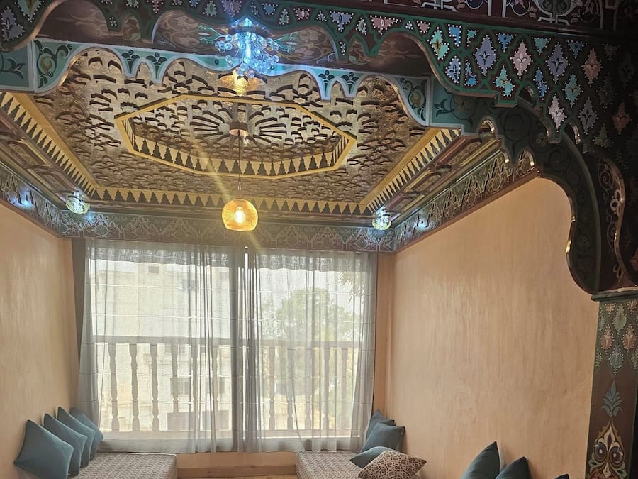 Ein Zimmer mit Holzboden, zwei Sofas mit Kissen, einem großen Fenster mit leichtem Vorhang und aufwendigen Verzierungen an Decke und Wand.