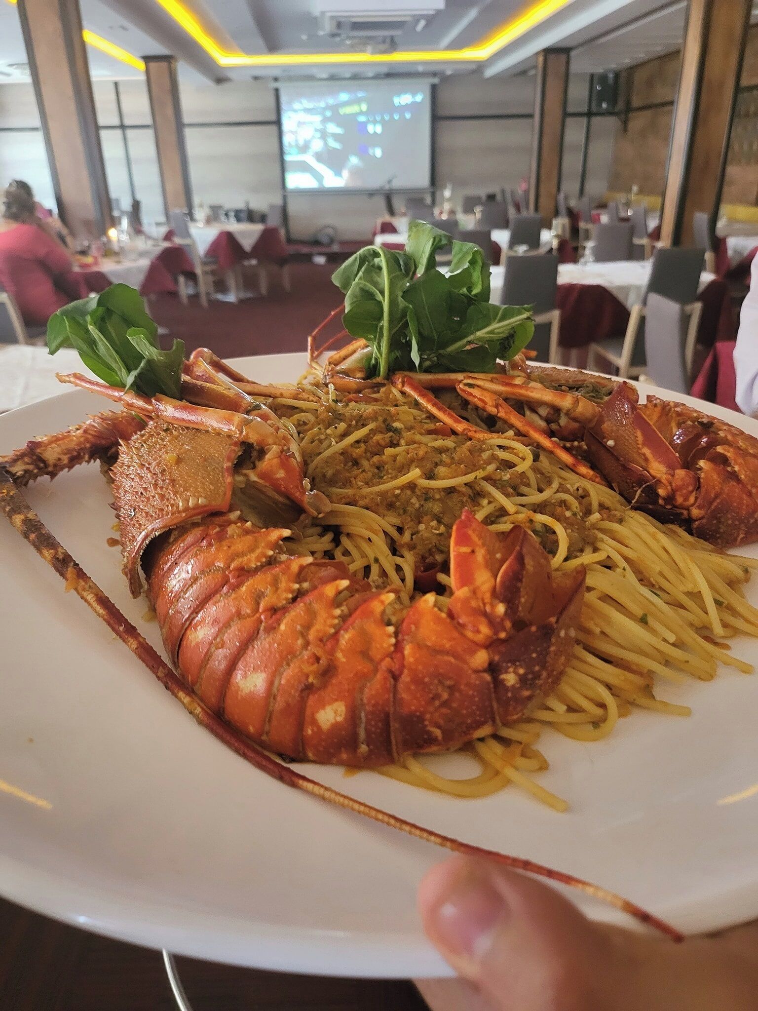 A lobster dish on a white plate with spaghetti and greens, in a restaurant with tables and chairs.